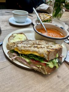 A grilled sourdough sandwich cut in half shows ham, tomato, greens, avocado, and melted cheddar, served with tomato soup on a wooden table.