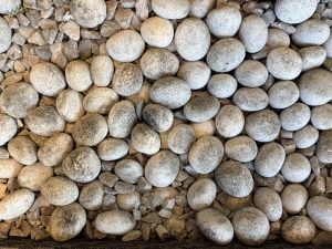 A close-up of smooth, grey pebbles interspersed with smaller, angular rocks, creating a textured pattern. The scene feels calm and natural.