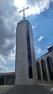 A low-angle shot highlighting the church's bell tower. The structure has a square footprint and is clad in gray and cream-toned stone slabs, featuring a vertical strip of dark glass running down the center of one of its faces. The tower culminates in a truncated pyramidal base, atop which stands a Latin cross.