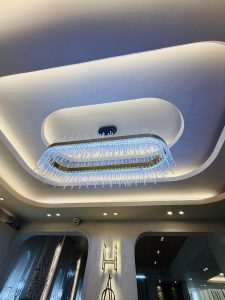 Modern chandelier with blue glass tubes hangs from an oval recessed ceiling, creating a luxurious ambiance. Soft lighting adds elegance and charm.