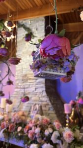 A beautifully decorated interior featuring hanging silk flowers in shades of pink and purple. In the background, a stone wall with a wooden ceiling provides a warm ambiance. 