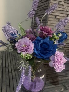 A vibrant arrangement of artificial flowers in a clear glass vase, featuring blue roses, soft purple blooms, and accented with lavender stems. 