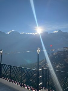 Snow-capped mountains rise beneath a clear blue sky, with the sun casting rays of light over the peaks. A black railing with decorative lamps frames the elevated viewpoint, overlooking a lush green valley below.
