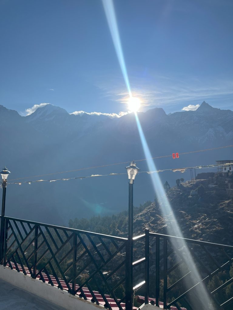 Snow-capped mountains rise beneath a clear blue sky, with the sun casting rays of light over the peaks. A black railing with decorative lamps frames the elevated viewpoint, overlooking a lush green valley below.