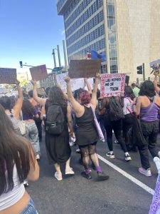A crowd of people participating in a feminist march on March 8, holding various signs with messages about social issues. The scene is lively with participants wearing purple clothing. 