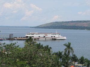 A large white public ferry with red and green stripes is docked on a calm body of water. Lush green palm trees and thick vegetation line the foreground, while a long, rolling hill stretches across the horizon under a pale blue sky. The scene depicts a quiet moment of transport and local scenery.
