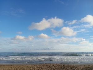 A peaceful beach scene with soft waves rolling toward the shore. The sky is bright blue with scattered fluffy clouds, creating a relaxing and open atmosphere. The horizon is clear, and the gentle motion of the ocean gives a sense of calm and tranquility.