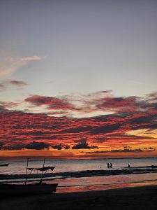 Fiery red and orange sunset clouds dominate the sky above a calm ocean, reflecting vibrant hues; silhouettes of two people and a structure on the sandy beach.