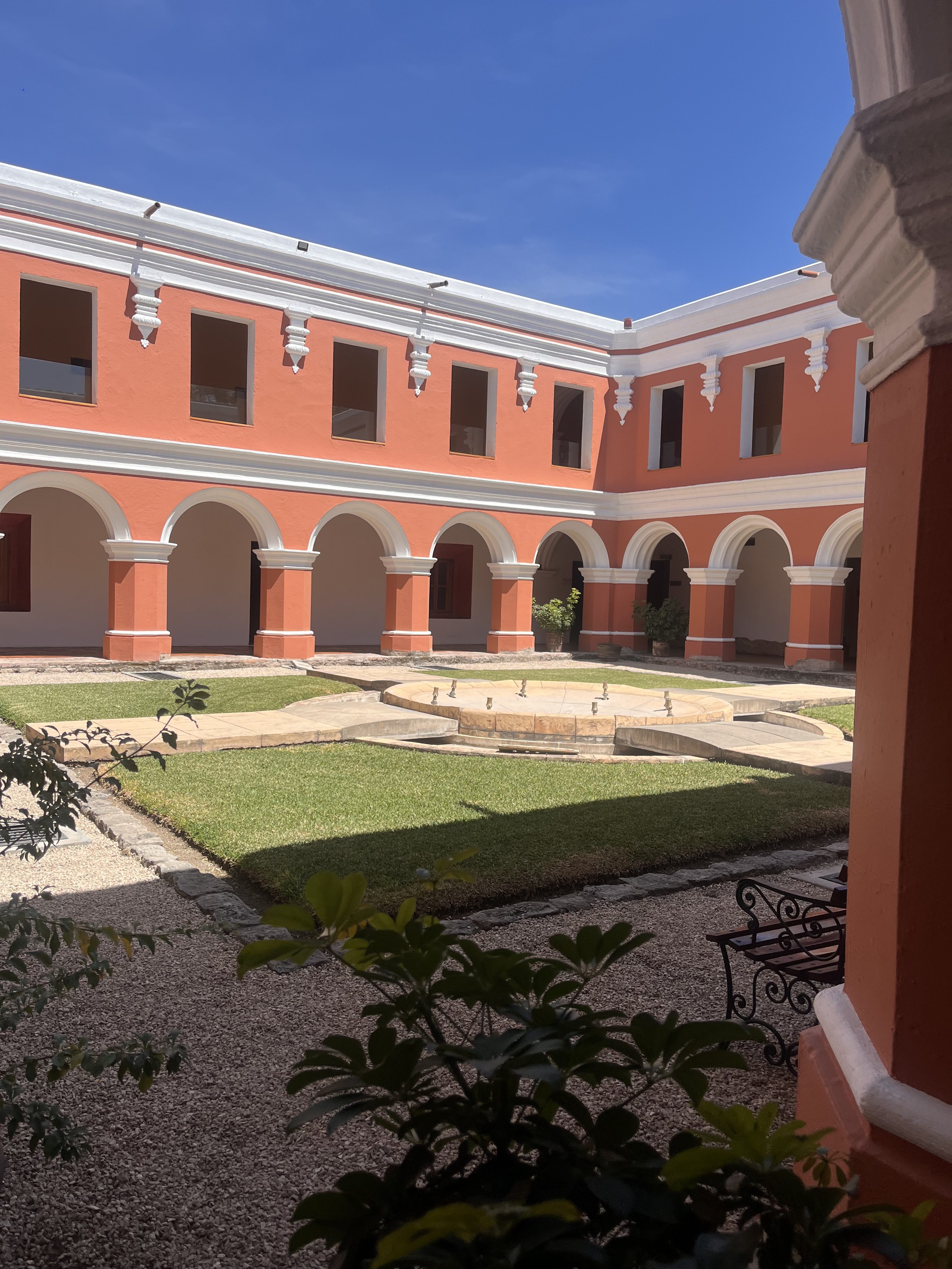 A courtyard with a circular stone fountain as the centerpiece, surrounded by lush green grass. The courtyard is enclosed by orange-painted buildings with white accents and arched doorways. The sky above is clear and blue, and the scene is partially framed by green foliage in the foreground.