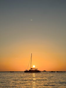 A tranquil sunset over the ocean, featuring a silhouetted sailboat with people on board