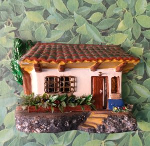 A small, colorful decorative figurine of a house is set against a leafy green background. The house has a tile roof with red and brown colors, yellow accents along the edges, and features a brown wooden door with a silver doorknob. There are two windows with decorative grilles and potted plants at the base, including leaves and small flowers. The figurine is detailed with a stone-like base, and a small staircase leads up to the entrance.