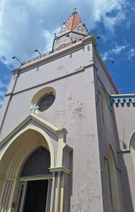 A close-up shot of a historic church featuring a tall, pointed steeple with a red and white roof. The building is predominantly pink with some areas showing signs of wear and peeling paint. Ornamental details can be seen above the entrance and windows, and a small cross is visible at the top of the steeple. The sky is bright with scattered clouds, providing a contrasting blue backdrop to the architecture.
