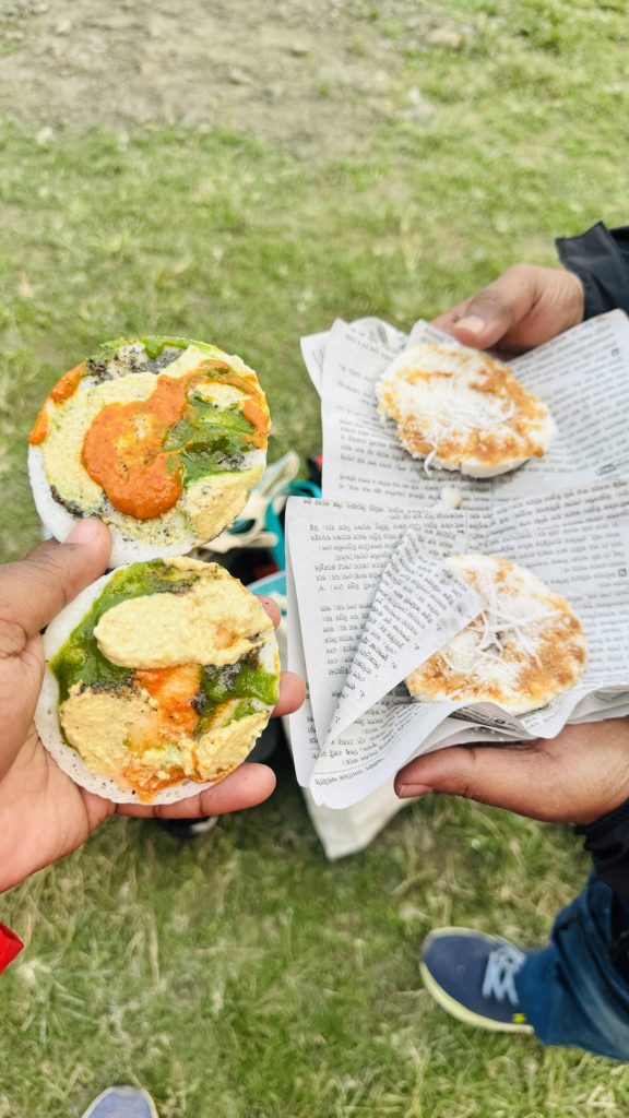 A first-person view of hands holding traditional rice cakes with chutneys and coconut in an open grassy field.