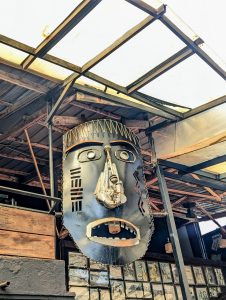 A large, stylized mask sculpture made from recycled metal and industrial parts is hanging from the ceiling in an industrial-style interior.
