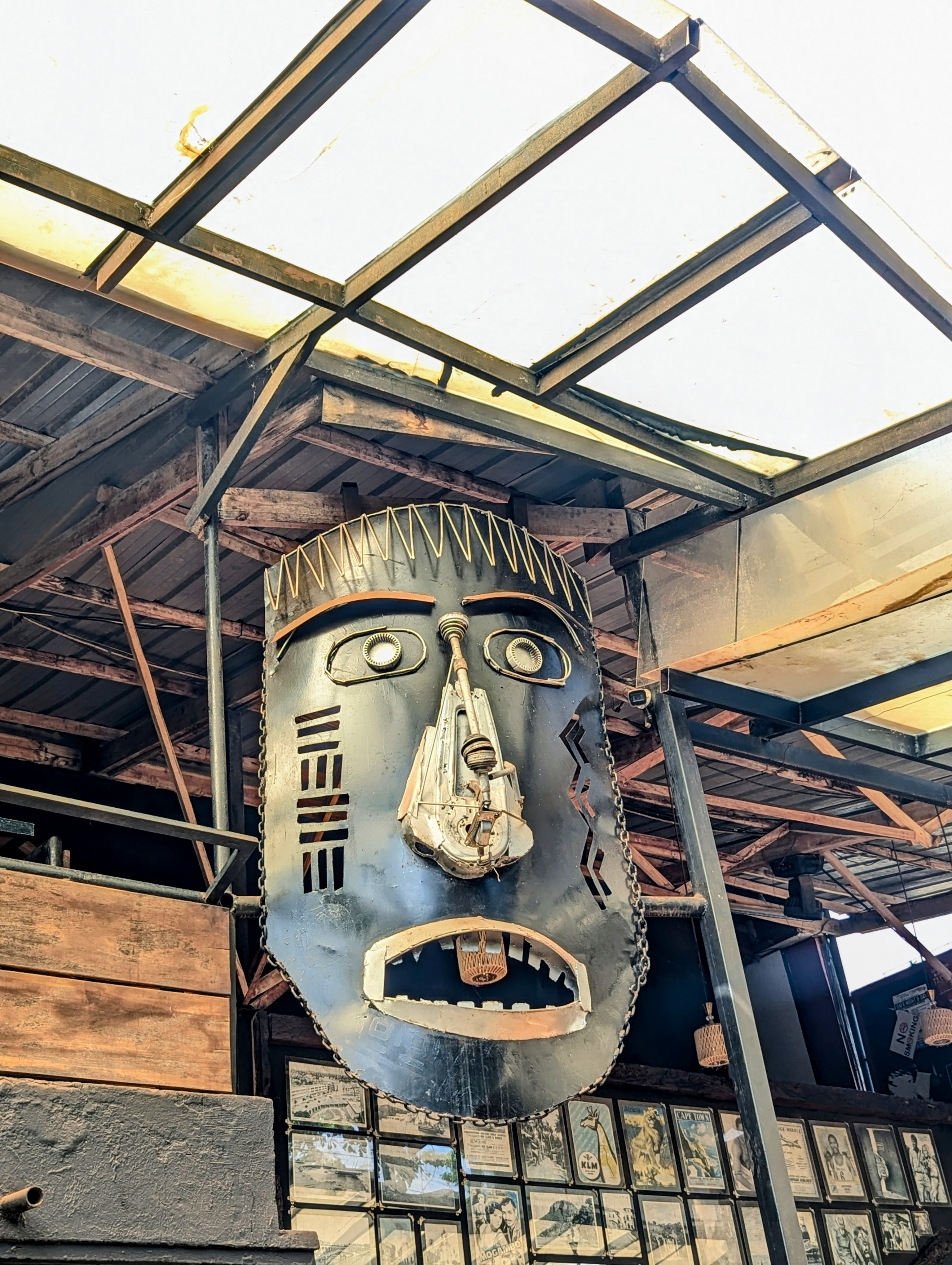 A large, stylized mask sculpture made from recycled metal and industrial parts is hanging from the ceiling in an industrial-style interior.
