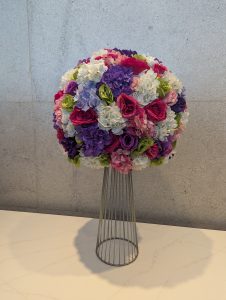 A round floral arrangement of pink roses, purple hydrangeas, and white flowers sits on a wire-frame pedestal against a gray wall, casting a soft shadow on a light countertop

