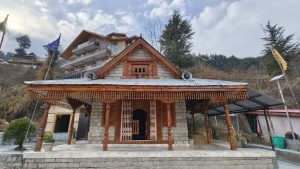 This photograph captures a serene Himalayan temple setting, likely located in a mountainous region of northern India, such as Himachal Pradesh or Uttarakhand, where traditional wooden architecture is deeply rooted in local culture. The temple in the foreground appears to follow the classic Kath-Kuni style, a construction technique that combines wood
