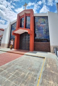The image shows the entrance of a modern church building featuring a prominent red and white façade. Above the entrance, there is a large cross. The church's front is adorned with colorful stained glass windows depicting religious figures. A pathway leading to the entrance is visible, with a decorative wooden cross and white cloth draped nearby. The sky is bright with scattered clouds, adding to the inviting atmosphere of the location.