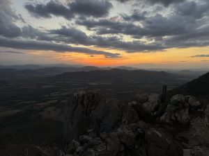 Sunset over mountain ranges with orange and yellow tones beneath a cloudy sky.