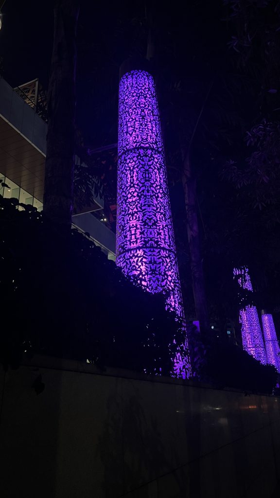 A nighttime scene featuring tall of fence of the Jio World Convention Center in Mumbai, India, illuminated columns with intricate patterns, bathed in a vibrant purple light. The columns are surrounded by dark foliage and are set against the backdrop of a building. The ambiance suggests a modern urban setting with decorative lighting. the
