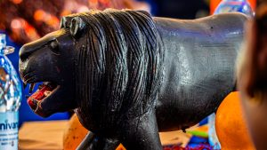 A side-view of a hand-carved lion sculpture made of dark polished wood, shown in a prowling stance with a textured mane and open mouth.