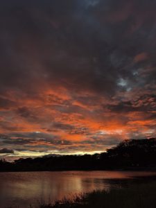 A dramatic sunset sky filled with deep orange and dark clouds above a calm lake, with soft reflections shimmering across the water