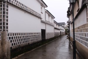 A narrow, wet street lined with traditional white buildings featuring black-and-white patterned walls in a Japanese setting.