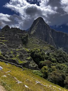 A panoramic view of Machu Picchu, showcasing ancient stone ruins perched on a steep hillside. The structures are set against lush green terraces and are surrounded by towering mountains under a partly cloudy sky. The foreground features grassy areas and some rocky terrain, while a few visitors can be seen exploring the site. The dramatic landscape reflects the beauty and historical significance of this iconic archeological site.
