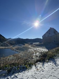 A clear blue sky dominates the scene with the sun shining brightly towards the upper right. In the foreground, a snowy landscape features patches of grass, a winding dirt path, and a small body of water to the left. A large, modern building with a slanted roof and several windows is situated on the right side, surrounded by a mountainous terrain that shows hints of snow on the peaks in the distance.