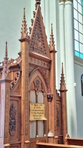 A carved wooden structure inside a church, featuring intricate designs and pointed spires. A wooden sign hangs on the structure, displaying a Spanish quote that translates to "If anyone is in Christ, he is a new creation; old things have passed away; all things have become new." The background includes neutral-colored walls and a glimpse of stained glass windows.
