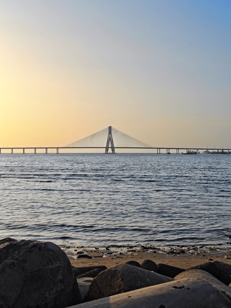 Sunset at Bandra-Worli Sea Link, with the bridge silhouetted against a warm sky and gentle waves along the rocky shore.