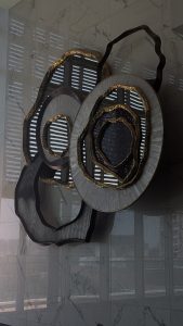 An abstract wall art piece with layered, irregular shapes and mixed textures, featuring metallic gold highlights and contrasting dark and light circular forms, mounted on a reflective marble wall with light and shadow patterns from nearby windows.