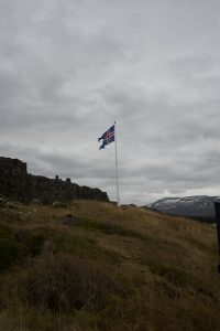 A flag with a blue background, featuring a red cross outlined in white, stands on a white pole amidst a grassy landscape. In the background, there are rocky formations and snow-capped mountains under a cloudy sky. The scene conveys a sense of open wilderness typical of Iceland's natural beauty.