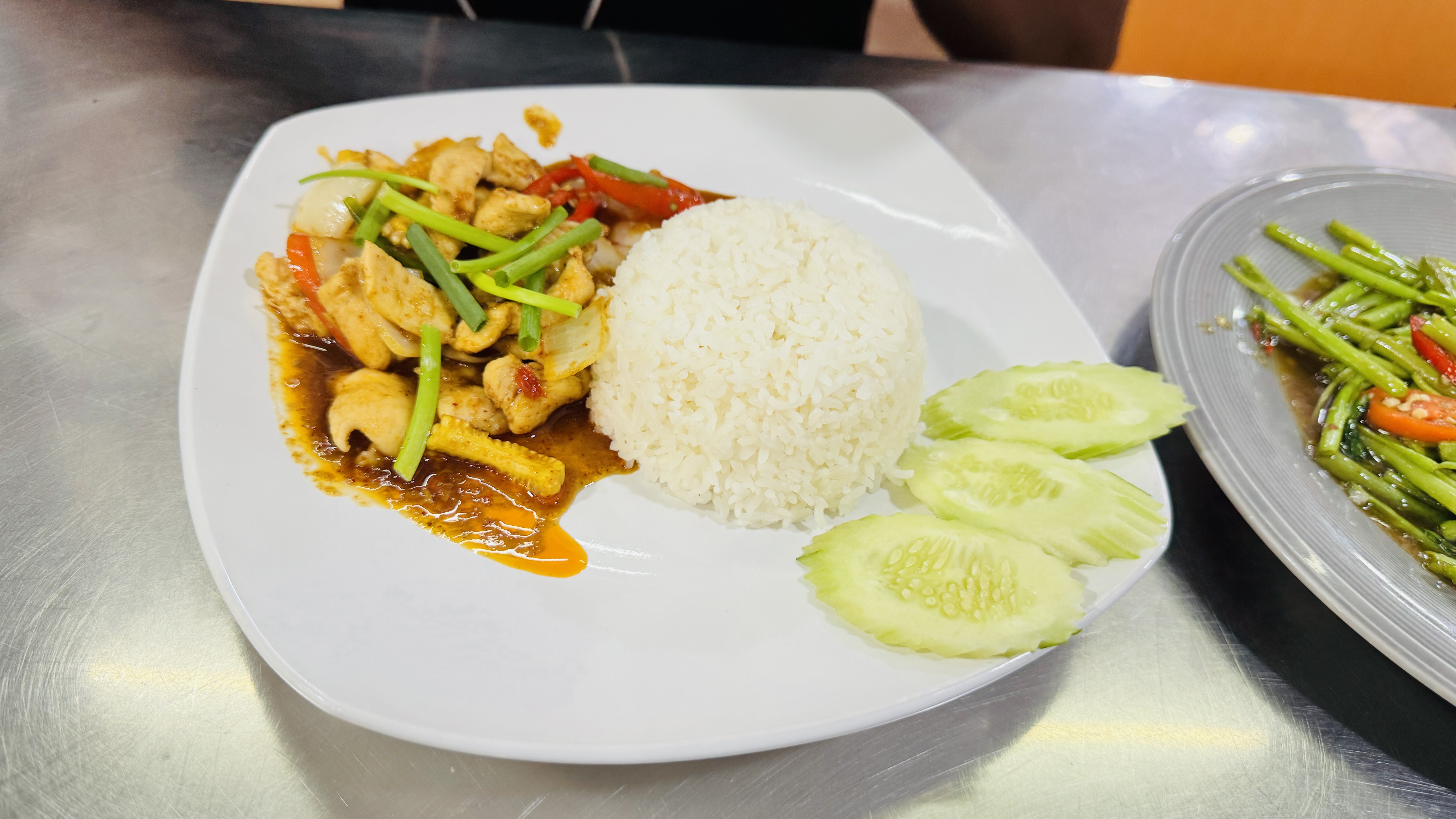 A white plate with steamed rice and chicken vegetable stir-fry in dark sauce, garnished with green onions, red peppers, and cucumber slices on a metal table.
