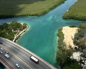 Aerial view of a turquoise waterway with mangroves, a roadside with vehicles, and a small landscaped park.