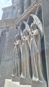 Three carved stone figures stand in a row, resembling angelic or religious figures. They are depicted with flowing robes and detailed facial features, set against a weathered stone backdrop that shows signs of aging. The figures are positioned prominently, with their heads slightly tilted and hands placed on their chests, giving an impression of reverence or contemplation. The scene is illuminated by soft sunlight, highlighting the texture and details of the carvings.