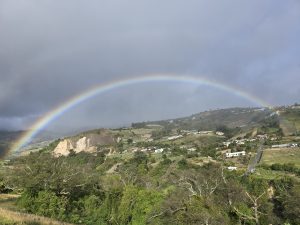 A wide-angle, panoramic landscape view of a perfect, vibrant rainbow stretching completely across a mountainous valley with scattered houses and fields in Costa Rica, under a dark, grey sky.