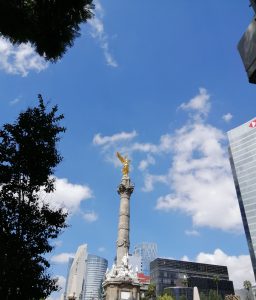 A tall Mexican Independence Angel monument with a golden eagle stands under a bright blue sky with white clouds.