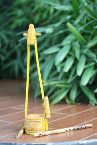 A traditional musical instrument made of wood and bamboo, featuring a yellow base with decorative carvings and a cylindrical resonator.
