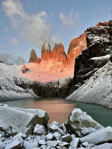Sunrise at the Torres del Paine National Park, with towering granite spires glowing orange over a teal glacial lake, snowy foreground rocks, and surrounding mountains.