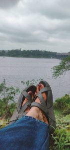A peaceful scene showing a person’s feet in sandals resting on grass beside a calm river. In the background are trees, distant buildings, and a bridge under cloudy skies.
