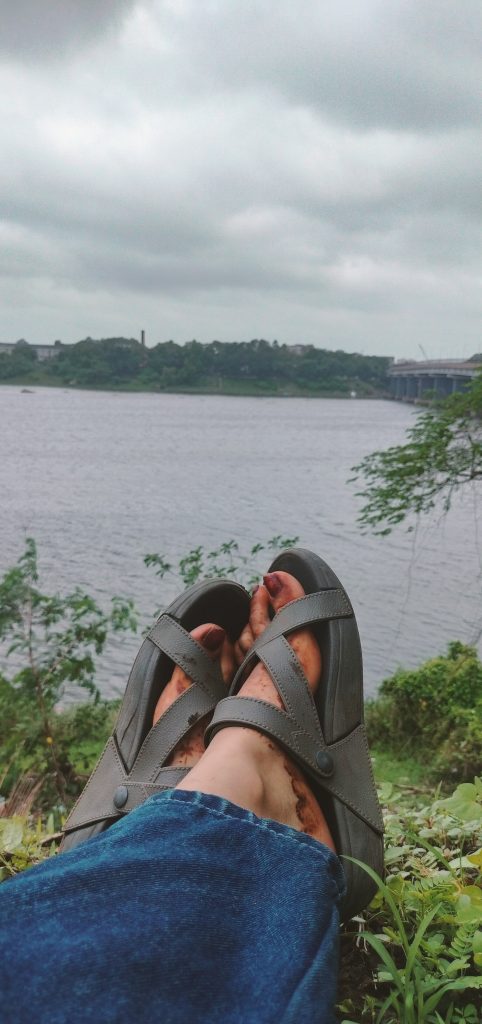 A peaceful scene showing a person’s feet in sandals resting on grass beside a calm river. In the background are trees, distant buildings, and a bridge under cloudy skies.