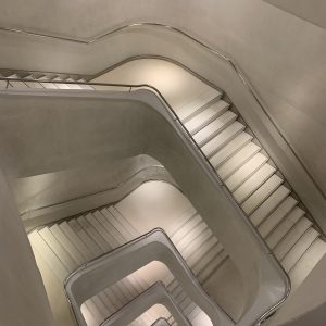 High-angle view looking down a modern, minimalist spiral staircase. Features smooth white concrete walls, white steps, and a continuous stainless steel handrail creating intricate geometric layers.