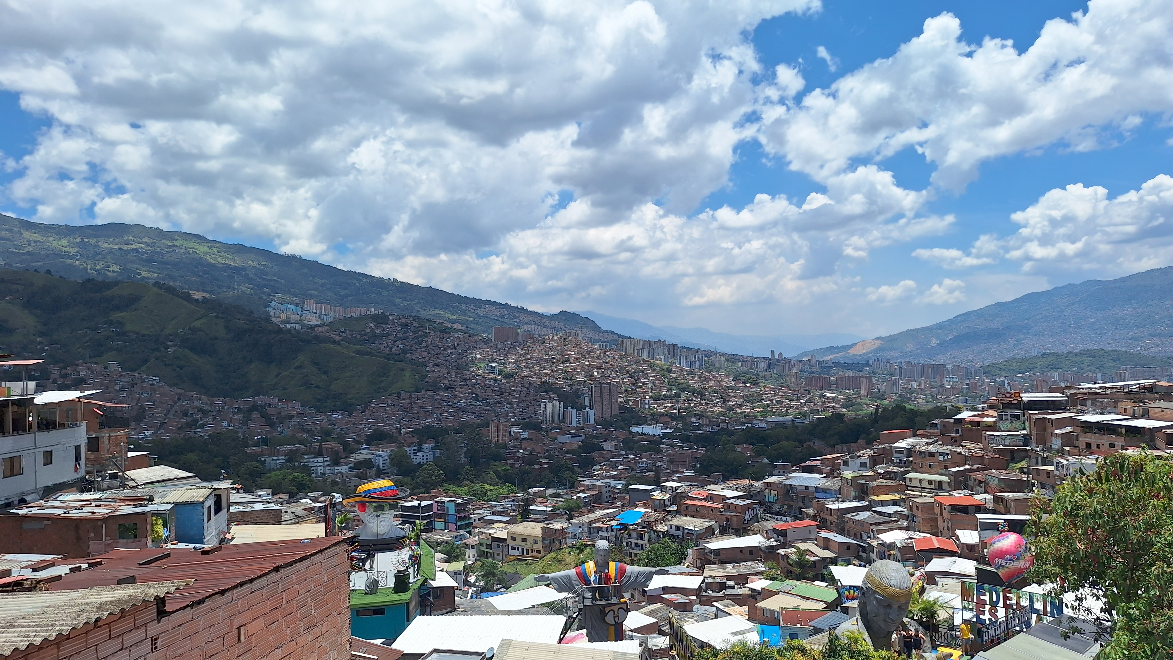 A panoramic view of Comuna 13 in Medellín, showing the dense cluster of brick houses climbing up the mountainside. The landscape features sculptures and urban decorations set against a blue sky dotted with thick clouds. The scene captures the vibrant life of the working-class neighborhoods that stretch out until they disappear into the valley’s hor