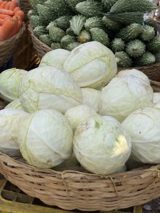 A woven basket filled with several heads of cabbage, showcasing their pale green color and crinkled leaves. In the background, there are baskets of carrots and a variety of bitter melons. The scene is set in a market environment.
