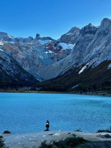 A lone hiker stands at the shore of Laguna Esmeralda, a glacier-fed lake near Ushuaia whose vivid turquoise water fills the frame. Behind, a steep glacial valley opens into jagged snow-patched peaks under a cloudless blue sky. Autumn-tinged lenga beech forest lines the right slope. Tierra del Fuego, Argentina.
