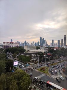 A cityscape view featuring a busy intersection with traffic, including cars and motorcycles, surrounded by lush trees. In the background, a skyline with tall modern buildings under a cloudy sky. A digital billboard is visible on one side of the road, and a mix of commercial and residential buildings is seen in the foreground.