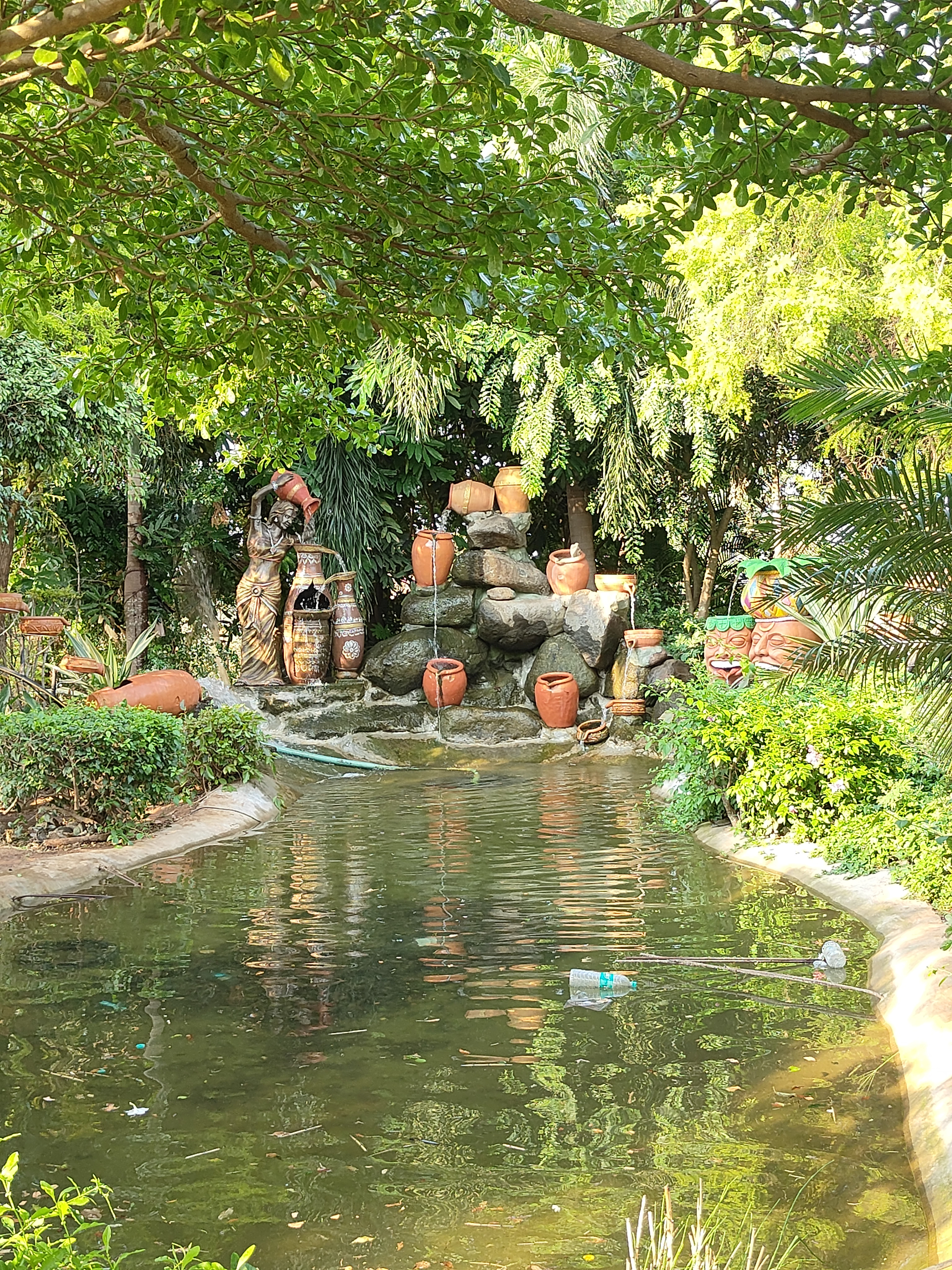 Serene garden pond surrounded by lush greenery, with decorative stones and cascading clay pots, and a statue of a woman carrying a pot.
