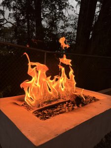 Intense orange flames burn in a modern fire pit set atop a square concrete base lined with stones. In the background, dark trees and a metal mesh fence are visible, creating a nighttime or twilight atmosphere in an outdoor setting.
