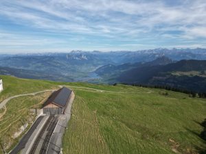 Mountain landscape in Switzerland with a lake in the valley, green hills, and a building with solar panels near a railway track.
